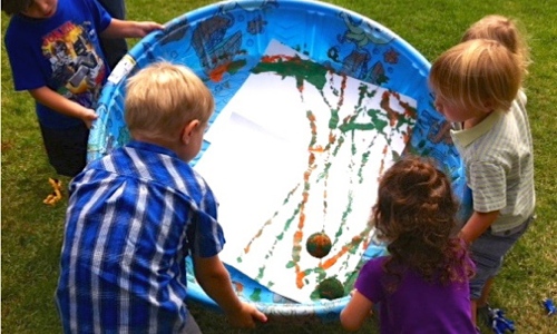 Kids using a tennis ball to paint on a large piece of paper in a kiddie pool