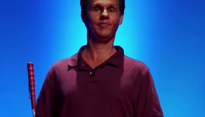 Daniel Kish in a collared shirt holding a cane against a blue background