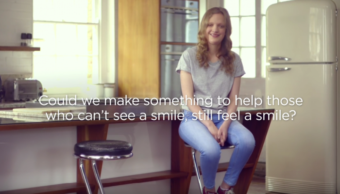 Woman sits on stool smiling. Text reads "Could we make something to help those who can't see a smile, still feel a smile?"