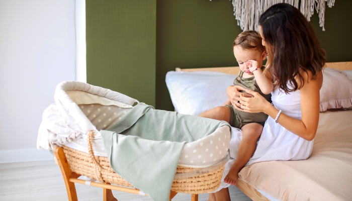 Mother with sleepy baby boy son sitting on bed near bassinet..