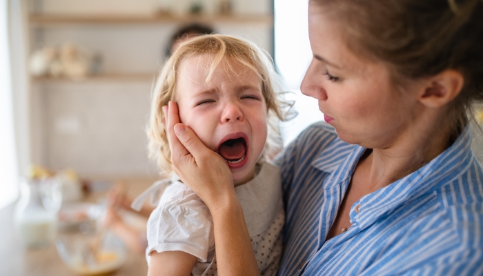 A mother holding a crying toddler daughter indoors in kitchen