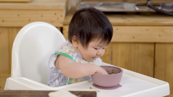 Baby sitting on a chair at home is grabbing food inside the bowl.