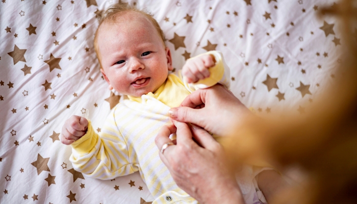 Cute newborn redhead baby in childish overalls lying crib during mother dressing.