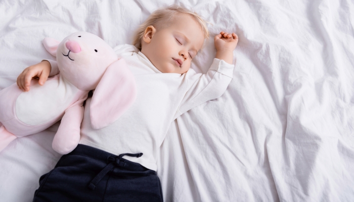 Overhead view of toddler boy sleeping on white bedding with toy bunny.