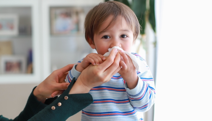 Adorable baby boy blowing nose into tissue paper by his mother at home