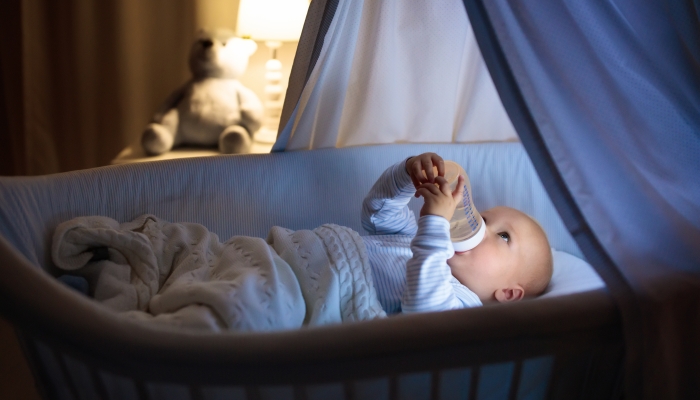 Adorable baby drinking milk in blue bassinet with canopy at night
