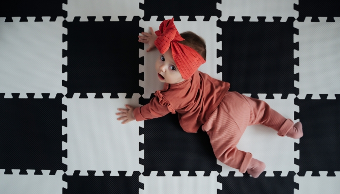 Adorable infant in stylish headband lying on puzzle mat at home.