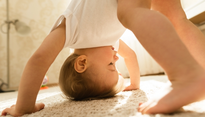 Cute playful little boy wearing bodysuit is standing upside down on the floor at his bedroom.