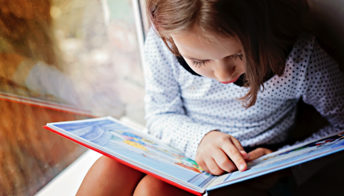 A little girl reading a book near a window.