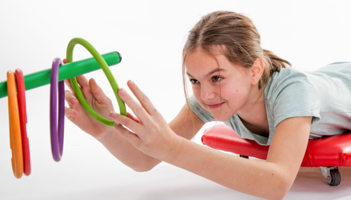 A girl on her belly on a scooter board reaches for rings.