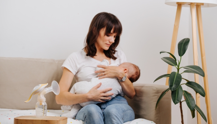 Mother is decently breastfeeding her baby sitting on a sofa with a breast pump on it.