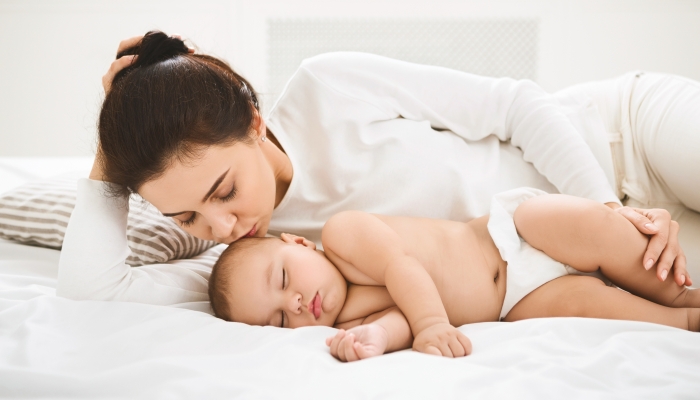 Young mother kissing her sleeping newborn baby in bed.