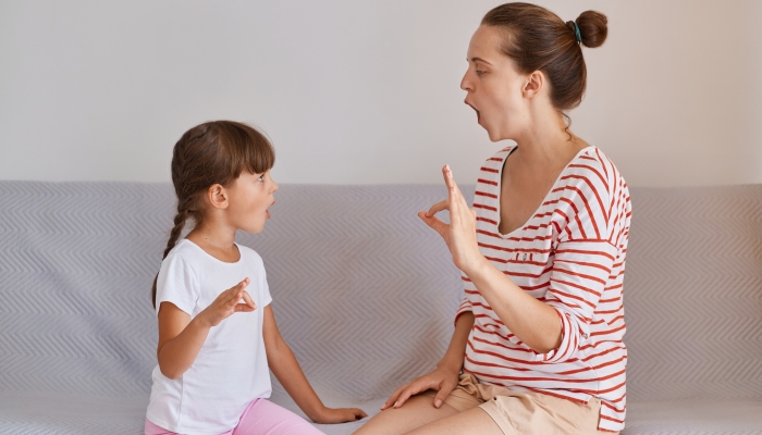 Female therapist sitting on sofa with little girl practicing correct pronunciation.