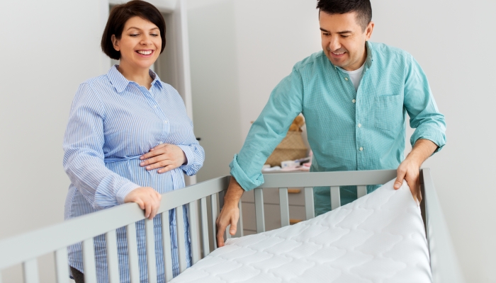 Happy middle-aged man and his pregnant wife arranging baby bed with mattress at home.