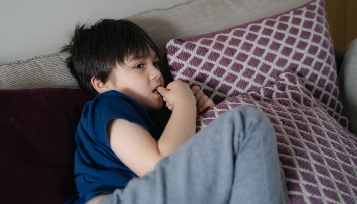 Kid lying on safa biting his finger nails while watching TV.