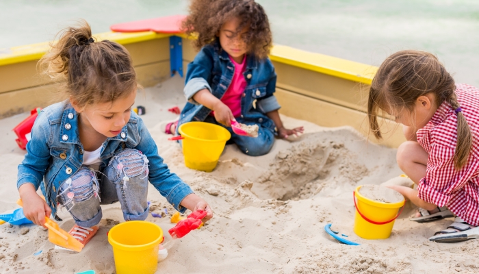 Three multiethnic little children playing with plastic scoops and buckets in sandbox at playground.