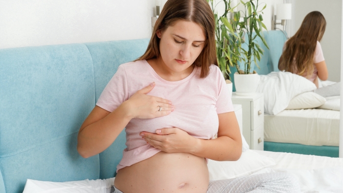 Young pregnant woman in pajamas sitting on bed and examining her breast.