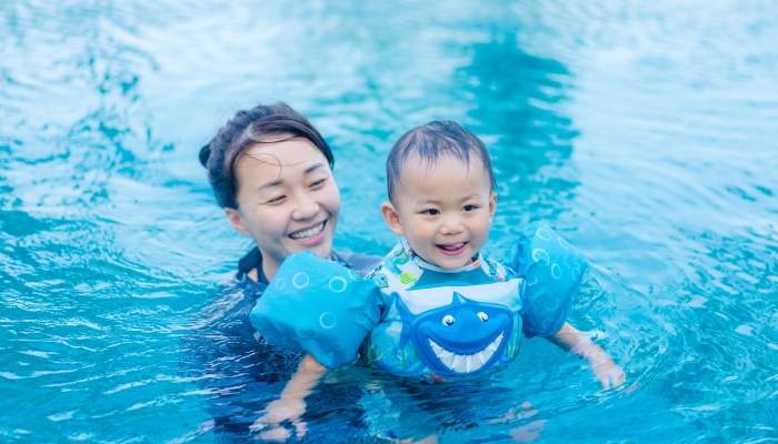 Asian mother and adorable little toddler boy with life vest having fun in a swimming pool.