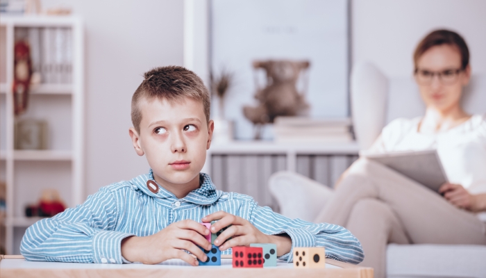 Autistic boy playing with colorful dice being watched by a psychotherapist sitting in the background.