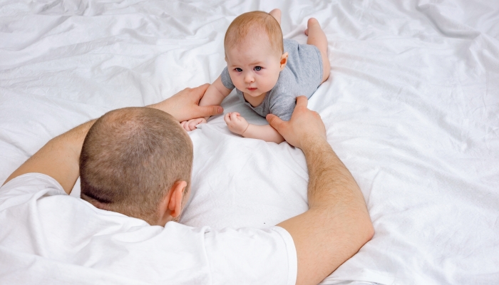 Father supporting baby laying on the bed teaching him to lay on tummy and hold his head up and roll over on tummy.