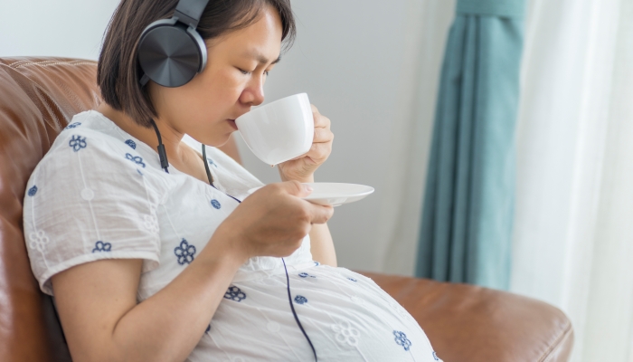 Pregnant woman sitting on a leather sofa enjoying music and black tea.