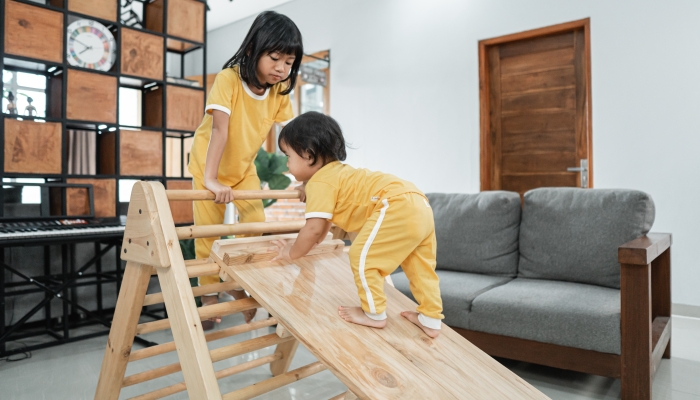 The baby climbs a pikler triangle toy accompanied by a sister in the living room.