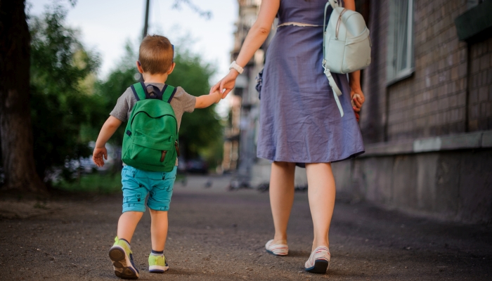 Back view of mother walking down the street with a little son with a backpack on summer day.