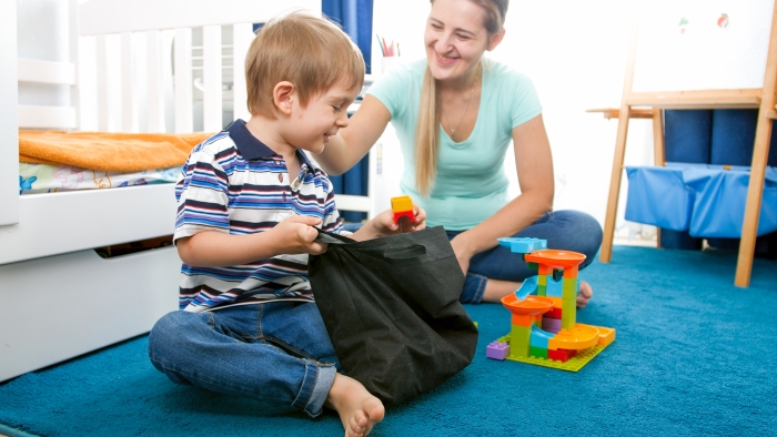 Cute smiling toddler boy tidying up his room and picking toys in bag.