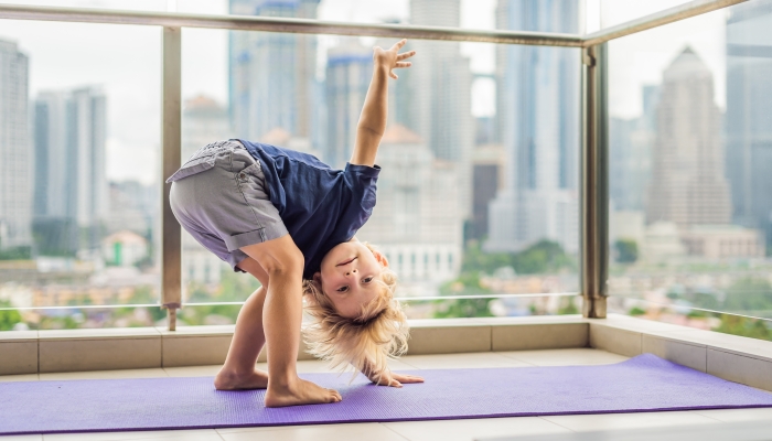 Happy little boy is practicing yoga on his balcony with a panoramic view of the big city.