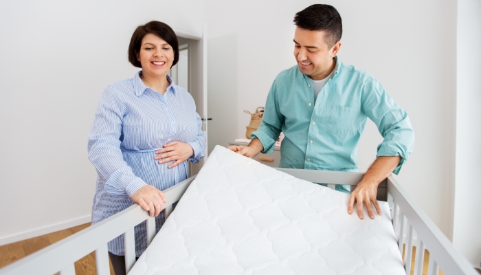 Middle-aged man and his pregnant wife arranging baby bed with mattress at home.