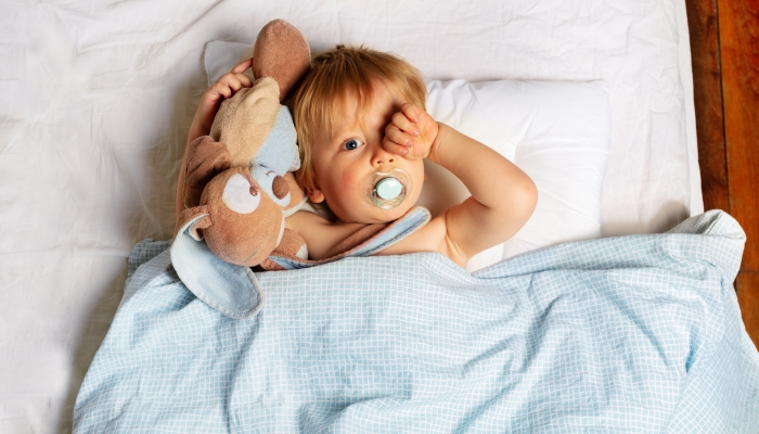 Sleeping baby boy waking up in the bed hugging toy and wipe eyes.