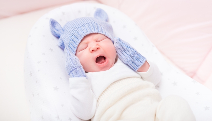 Yawning little baby wearing knitted blue hat with ears and mittens lying in beautiful cradle.