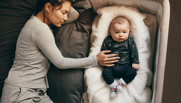 Young mother sleeping on bed with her infant baby sleeping on a bedside bassinet.