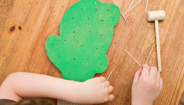 Little boy using a play hammer to stick toothpicks in a cardboard cactus.