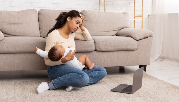Exhausted Black Mother Holding Crying Baby Sitting At Laptop Indoor.