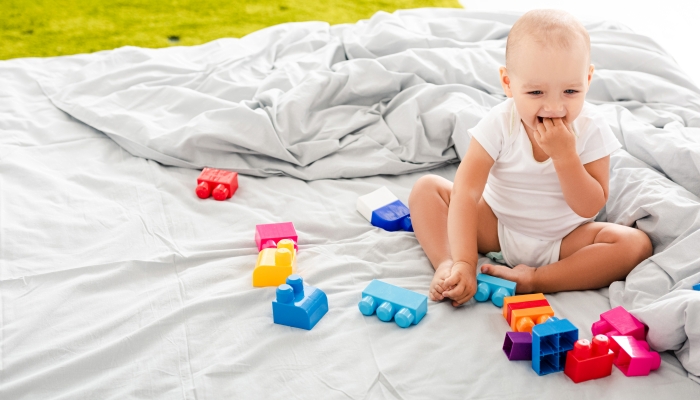 Funny barefoot baby in white clothes sitting on bed near colorful construction and taking fingers into his mouth.