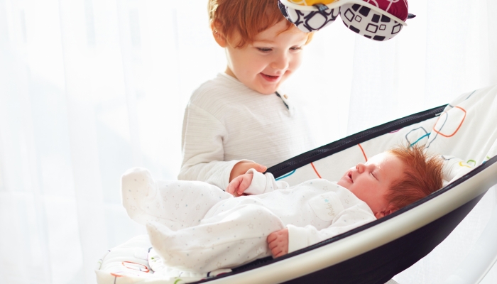 Happy toddler baby brother welcoming his little baby sister, that lying in cradle swings.