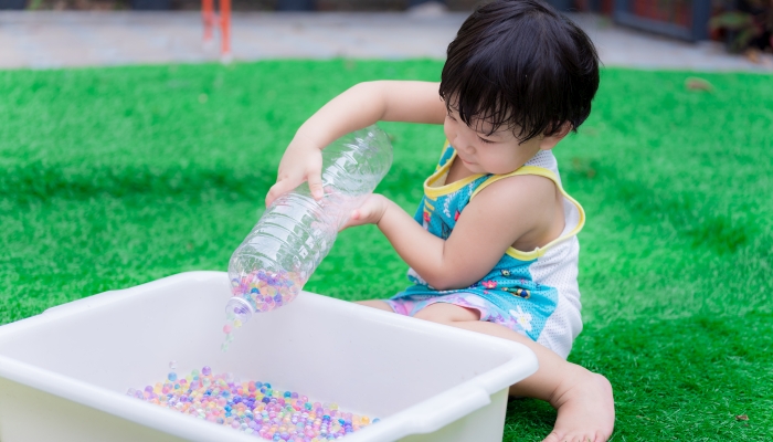 Preschool baby boy is using two hands to hold clear bottle to pour water beads.