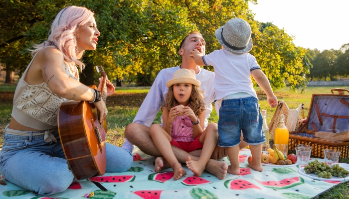 Family of four with mom playing guitar sitting picnicking in the park.