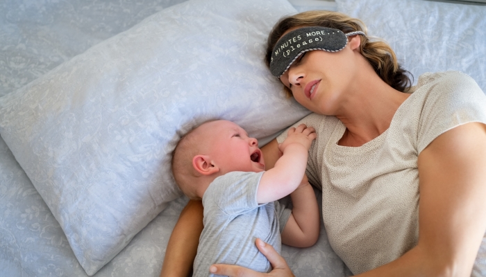 Tired mother with eye band trying to sleep while little toddler crying on bed.