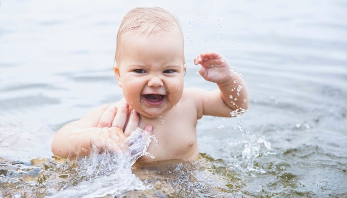 Beautiful baby bathes splashing water and laughing and mom supports daughter by hand.