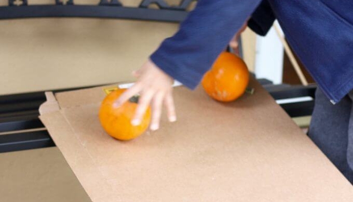 Child rolling pumpkin and orange down a cardboard ramp.