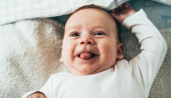 Close Up Photo of Adorable Baby Sticking Out his TongueFace of curious newborn baby boy looking at the camera with with tongue sticking out while lying in bed at home.