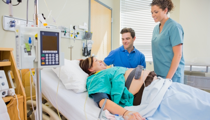 Happy nurse and man looking at pregnant woman lying on bed in hospital.