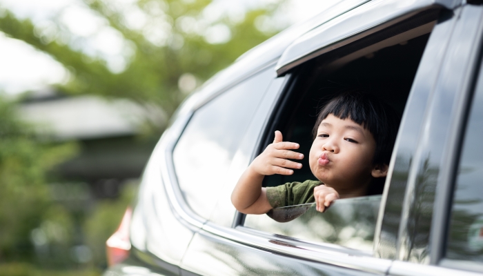 Happy asian boy waving hands gesturing hello out of the car window during a trip with his family.