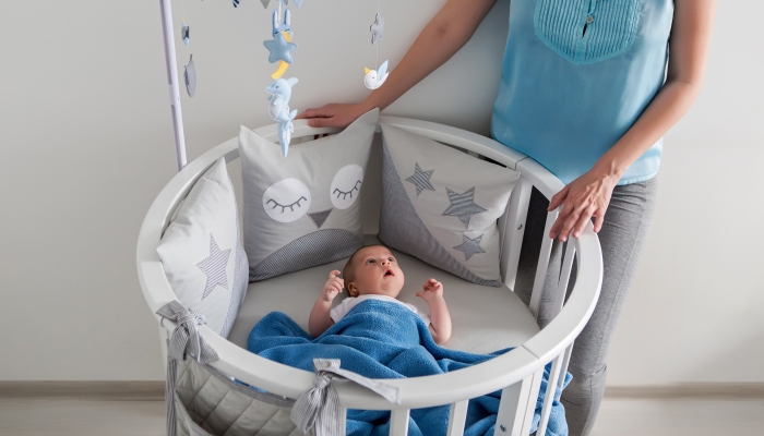 Mother in a blue jacket stands next to a round white crib for a newborn baby.