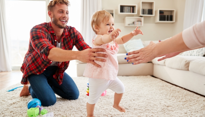 Toddler girl walking from dad to mothers arms in sitting room.