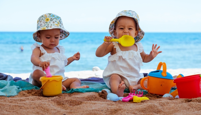 Twin baby girls sitting on a beach by the sea playing with sand toys wearing white bodies and colorful bucket hats.