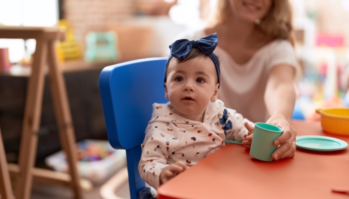 Woman and toddler learning to eat with plastic dish toy sitting on table at kindergarten.