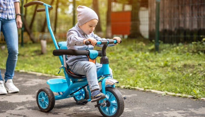 Little boy is learning to ride a children's tricycle while walking with his mother.